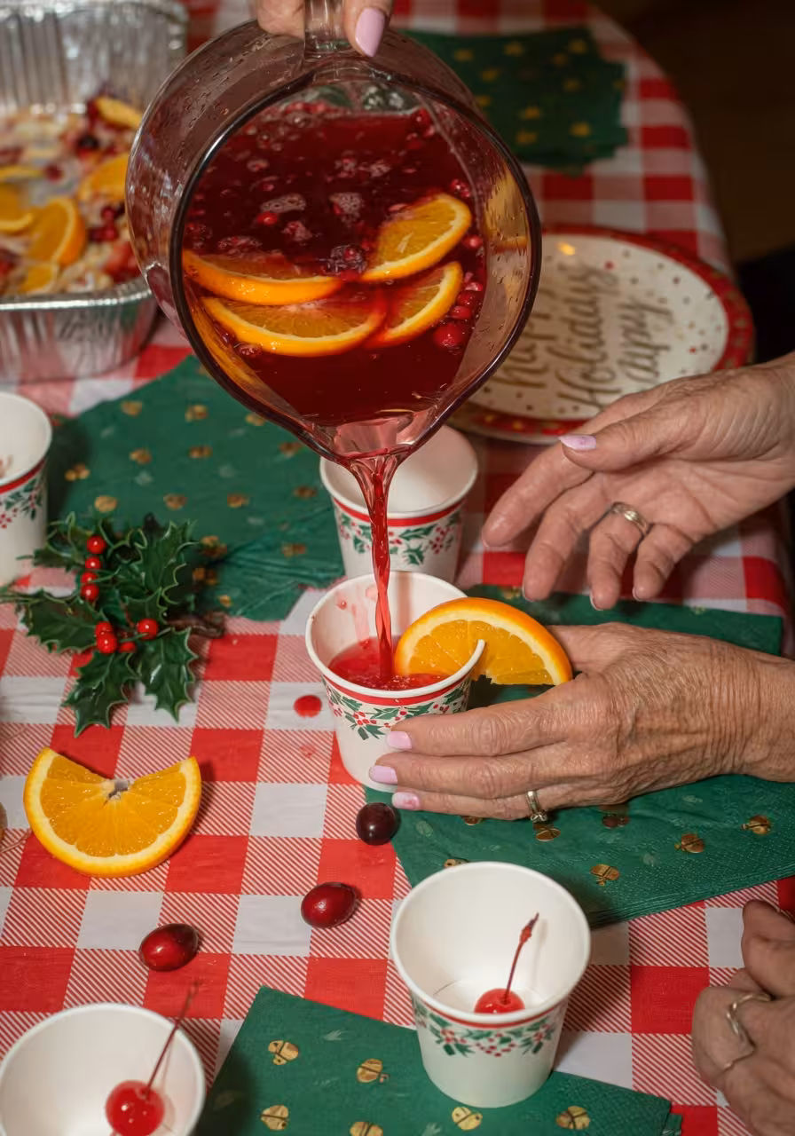 Close-up of holiday punch being poured into small paper cups, cranberry-red liquid with orange slices visible